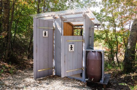 Holzgebäude mit zwei Toilettenkabinen und einem großen Behälter daneben, umgeben von Bäumen.