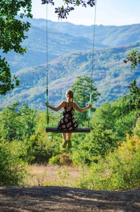 Junge Frau auf einer Schaukel mit Blick auf bewaldete Berge und grüne Landschaft.