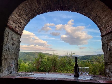 Blick durch einen Steinbogen auf eine malerische Landschaft mit Weinflasche und Gläsern.