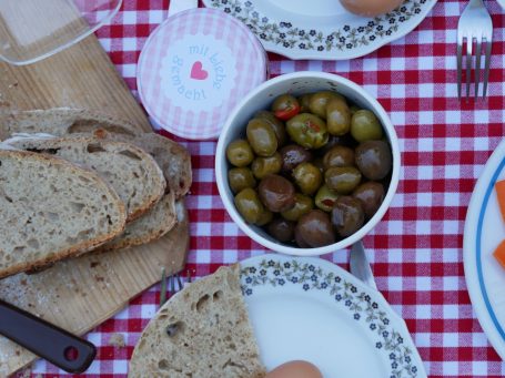 Ein Tischgedeck mit Brot, Oliven, einem gekochten Ei und Obst auf kariertem Tischtuch.