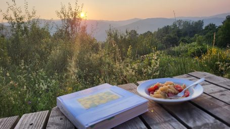 Ein Tisch mit einem Buch und einem Teller Essen, Blick auf eine Landschaft bei Sonnenuntergang.