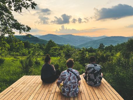 Drei Personen sitzen auf einer Holzplattform und schauen auf die bergige Landschaft bei Sonnenuntergang.